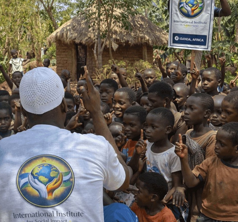 A member of the Institute team, wearing a white shirt with the Institute logo, speaking to a crowd of children in front of a thatched home in a Ugandan village